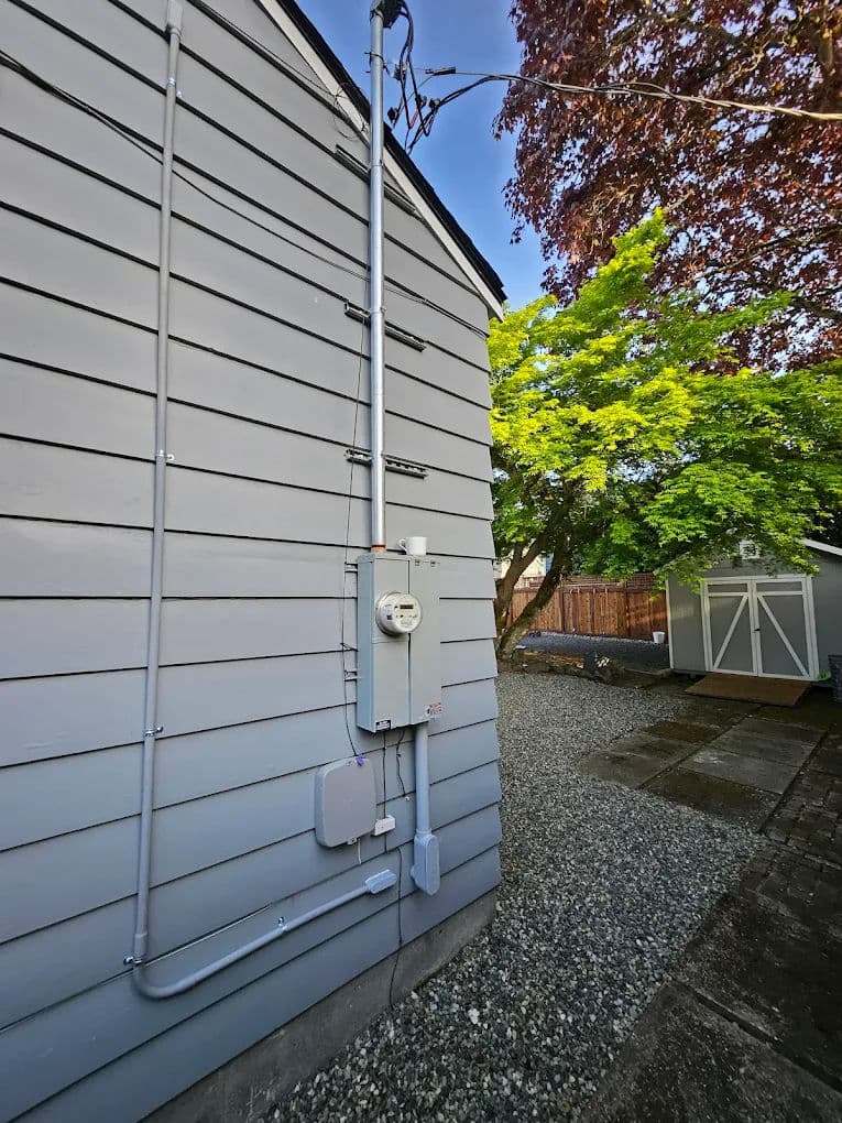 Electric meter and service panel installed on gray house siding with trees in the background.